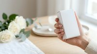 Young woman with medium skin tone holding blank white box with elegant table setting and white roses in background, suitable for product packaging mockups and lifestyle marketing.