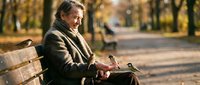 Mature man with gray hair and stubble, wearing brown jacket and scarf, sitting on park bench reading newspaper in autumn setting with golden foliage.