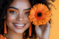 Young African American woman with bright smile and curly hair holding orange gerbera daisy, wearing matching orange earrings. Natural beauty and joy concept.