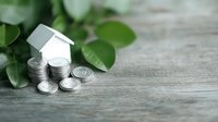 Small white paper house model with stack of silver coins on wooden surface with green plant leaves for real estate investment concept.