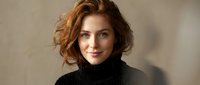Young Caucasian woman with curly red hair wearing black turtleneck sweater smiling warmly at camera against neutral beige background for lifestyle portrait.