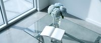 Modern glass coffee table with white hydrangea flowers in vase and open book, minimalist interior design for contemporary living room decor.
