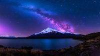 Majestic snow-capped mountain peak reflected in calm lake waters under vibrant purple and blue night sky with bright Milky Way galaxy stretching across horizon.