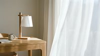 Wooden desk with table lamp and white curtains in bright, minimalist home office or bedroom interior space.