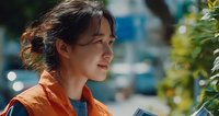 Young Asian woman in orange jacket smiling outdoors with blurred urban background and greenery, conveying happiness and positivity in natural daylight.