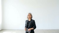 Middle-aged woman with gray hair and light skin tone wearing professional black blazer and black top, standing confidently in minimalist white interior space for corporate business portraits.