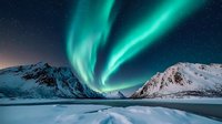 Spectacular aurora borealis dancing over snow-covered mountain peaks and frozen lake landscape under starry night sky creates dramatic natural light show in polar region.