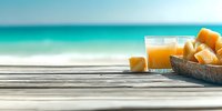 Fresh tropical mango juice in glass with cut fruit pieces on wooden table at beach, turquoise ocean background, summer refreshment concept.
