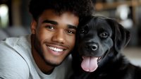 Young African American man with bright smile embracing black Labrador dog, both looking at camera with joyful expressions in cozy home setting.