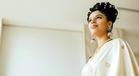 Young woman with dark hair styled in an updo, wearing pearl earrings and a white draped garment, gazing upward in an elegant interior setting with neutral tones.