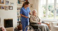 Female healthcare worker in blue scrubs assisting smiling elderly Caucasian woman in wheelchair at bright home interior with photo gallery wall.