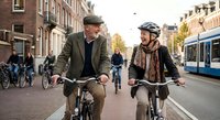 Senior couple cycling together through European city street during golden hour sunset with tram and historic architecture in background.