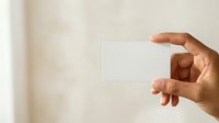 Person of medium skin tone holding blank white business card or paper against neutral beige background, suitable for corporate branding, marketing mockups, and professional presentations.