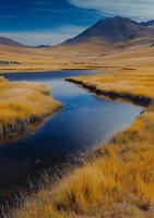 Winding river through golden grasslands beneath majestic mountain peaks, reflecting deep blue sky in calm waters. Dramatic wilderness landscape for travel and nature themes.