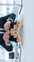 Businessman in suit arranging wooden blocks with business icons on white desk in modern office environment for strategic planning and development.