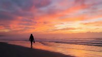 Silhouette of female surfer holding surfboard standing on sandy beach at golden hour sunset with vibrant orange and pink sky, ocean waves, and coastal landscape for travel and lifestyle content.