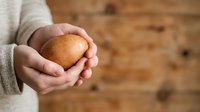 Person with light skin tone holding a fresh round bread roll in their hands against a wooden background, suitable for bakery, food, and culinary content.