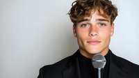 Young Caucasian male performer in black attire holding microphone against light background, expressing confidence and charisma for entertainment and talent concepts.