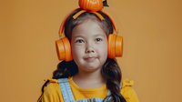 Young Asian girl wearing bright orange headphones and yellow sweater smiling at camera against orange background. Studio portrait capturing childhood joy and modern technology.