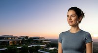 Young woman with dark hair and olive complexion wearing casual gray t-shirt, standing outdoors at sunset overlooking modern suburban development and rolling hills, suitable for lifestyle and real estate marketing.