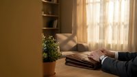 Businessman hands stacking folded clothes on wooden table in warm sunlit room with bookshelf and plant creating cozy domestic atmosphere for organization.