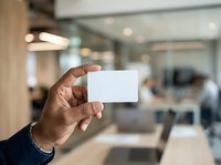 Hand holding blank white business card in modern office environment with blurred colleagues working in background, suitable for corporate branding and networking concepts.