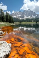 Dramatic mountain lake landscape with vibrant orange mineral deposits along shoreline, snow-capped peaks and evergreen forest reflected in calm waters. Suitable for nature prints.