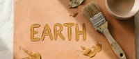 Young female artist with light skin tone working on clay sculpture, carving the word "EARTH" into terracotta surface with paintbrush and wooden tools nearby, overhead view of creative workspace.