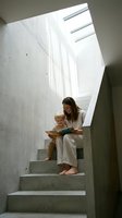 Mother and young child reading book together on concrete stairs in modern home with natural skylight creating dramatic shadows and peaceful atmosphere.