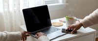 Two people working together at a home desk with a laptop, smartphone, and steaming tea cup near a bright window.