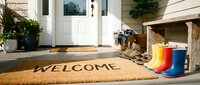 Sunny front porch with a welcome doormat, colorful rain boots, and rustic bench by a white door, evoking a warm, inviting home entrance.