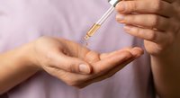 Woman applying golden serum oil from dropper bottle onto palm of hand for skincare routine and beauty treatment on purple background.