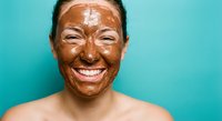 Happy Asian woman with chocolate face mask smiling at camera against turquoise background for skincare and beauty treatment concepts.