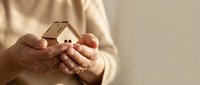 Elderly hands gently holding small wooden house model symbolizing home protection, real estate investment, mortgage planning and property security concept.