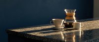 Ceramic cup and glass coffee maker on granite countertop with warm sunlight, suitable for café, kitchen, and beverage marketing materials.