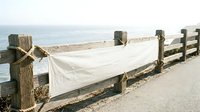 White fabric banner hanging on wooden fence posts along coastal pathway with ocean view and sandy beach in background.