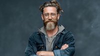 Smiling bearded middle-aged Caucasian man with glasses and curly hair in denim jacket and hoodie with crossed arms against dark background.