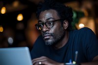 African American mature man in glasses working on laptop at night, focused expression while using computer in dark room with bokeh lights background.