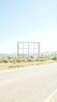 Blank billboard frame beside a sunny rural road with a Mediterranean village and olive trees in the background, copy space for advertising.