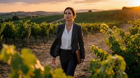 Professional businesswoman walking through vineyard at golden hour sunset with rolling hills and grapevines creating inspiring agricultural landscape scene.