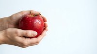 Fresh red apple with water droplets held in human hand against white background, representing healthy eating and nutrition.