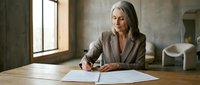 Mature Caucasian businesswoman in beige blazer writing documents at wooden table in modern office with natural lighting for corporate presentations.