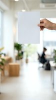 Hand holding blank white paper sheet in modern office environment with blurred background, suitable for mockups, presentations, advertising campaigns, and corporate branding materials.