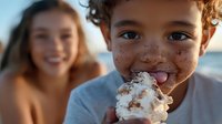 Young child with freckles enjoying ice cream at the beach, smiling with messy face while a girl blurs in background during summer vacation.