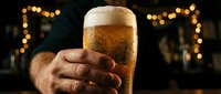 Hand holding a cold beer glass with golden foam head against blurred warm bokeh lights in dark bar setting, perfect for beverage, pub, and hospitality marketing.