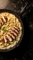 Creamy chicken alfredo pasta with grilled sliced chicken breast served in cast iron skillet on dark granite surface for restaurant menu and cooking content.