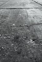 Cracked concrete surface with rough texture and debris photographed in dramatic black and white, showing urban decay and weathered patterns.