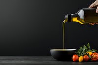 Pouring golden olive oil from dark bottle into black ceramic bowl with fresh tomatoes and herbs on wooden table against dark background.