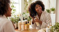 Young African American woman in bathrobe applying skincare products while looking in bathroom mirror, surrounded by natural plants and beauty bottles for morning routine.