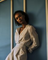 Young African American woman in elegant white wrap dress with puff sleeves poses against blue wall in moody natural lighting, creating atmospheric fashion portrait.